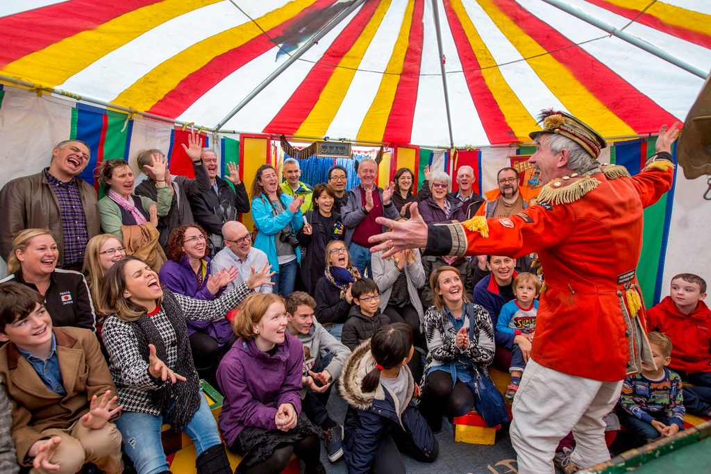 A man in circus costume performing to an audience in a colourful tent