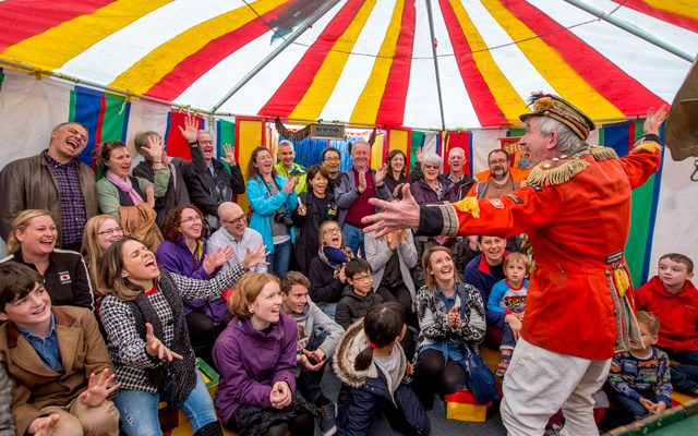 A man in circus costume performing to an audience in a colourful tent