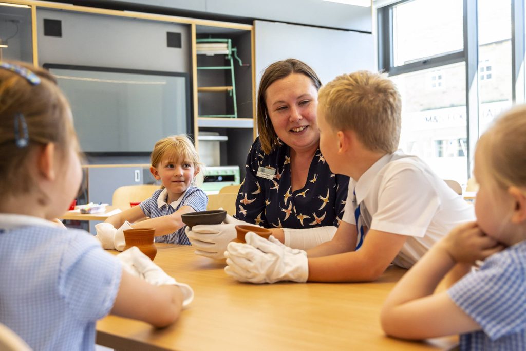 A woman is showing a museum object to some school children. They are all sat round a table and wearing white gloves.