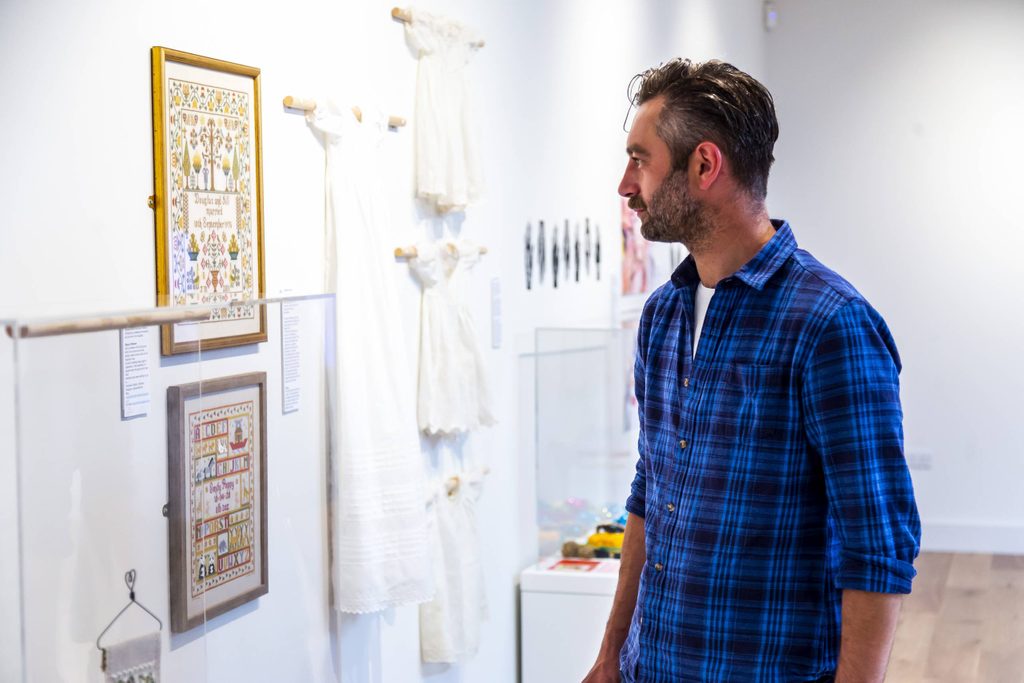 Man with short dark hair and a blue and black checked shirt looking at a framed embroidery picture in the exhibition gallery.