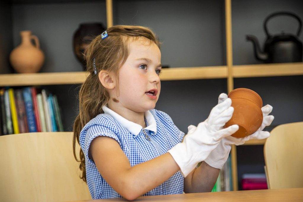 Young girl wearing a blue gingham school uniform dress is wearing white gloves and handling a clay pot.