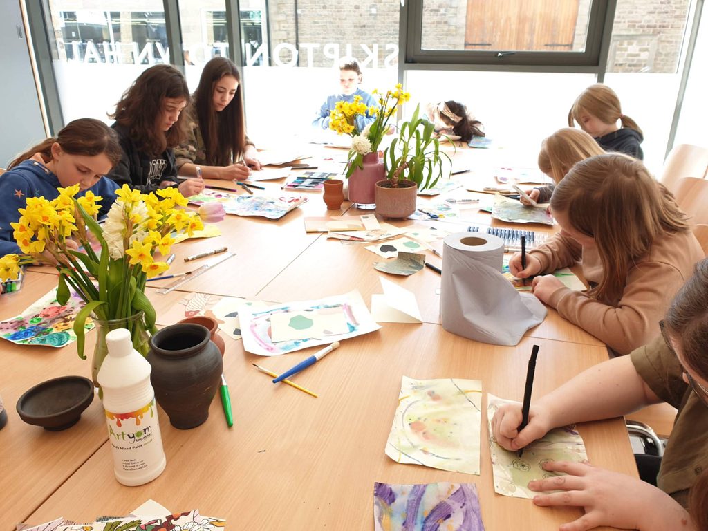 A group pf young people sit round a table . they are drawing.